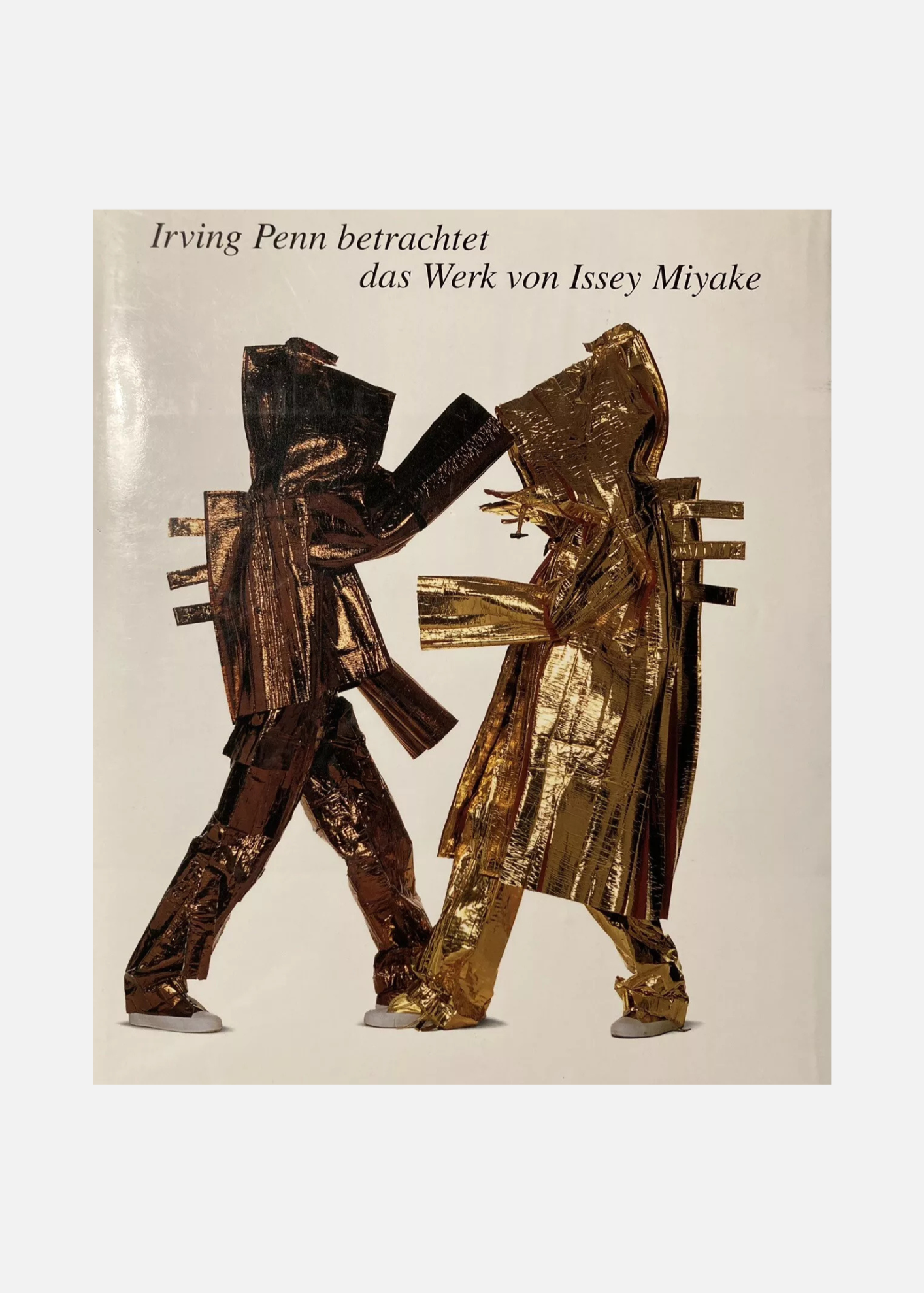 Couverture du livre "Irving Penn betrachtet das Werk von Issey Miyake", présentant deux figures stylisées en mouvement, vêtues de vêtements en matières dorées et texturées. Les silhouettes sont placées sur un fond beige, et les personnages semblent interagir de manière dynamique, mettant en valeur l'esthétique innovante des créations de Miyake. Le titre en haut est inscrit en lettres noires.