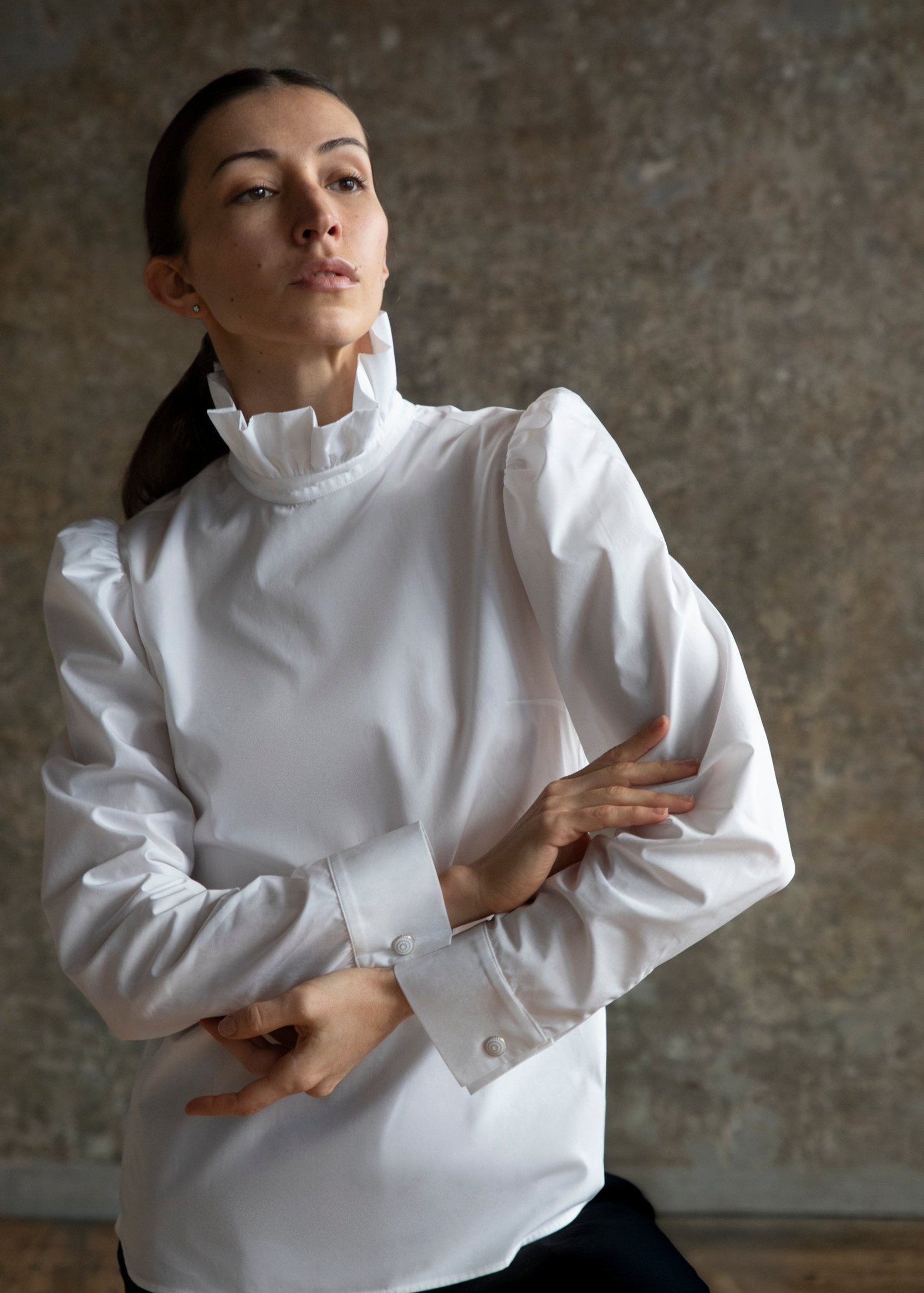 Une femme portant le Chemisier Sidonie, une blouse blanche à col haut et manches bouffantes, se tient devant un mur texturé gris, incarnant une élégance raffinée. Les bras croisés, parée de sublimes boutons de manchette, elle regarde au loin avec une expression contemplative.