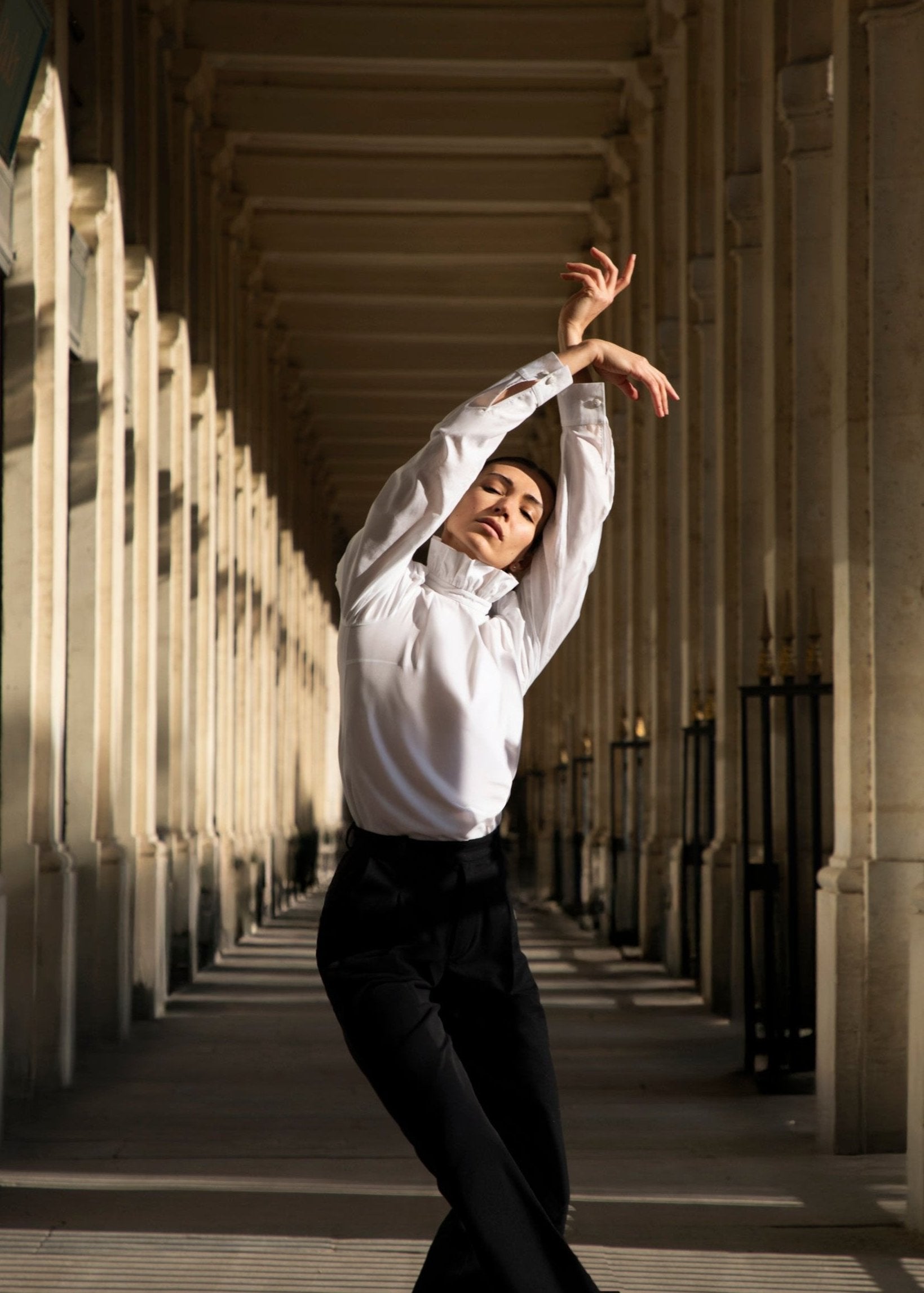 Une danseuse portant le chemisier Sidonie et un pantalon noir exécute gracieusement une pose artistique inspirée du ballet dans un couloir architectural, avec des colonnes projetant de longues ombres sur le sol, ajoutant une touche de luxe à la scène.