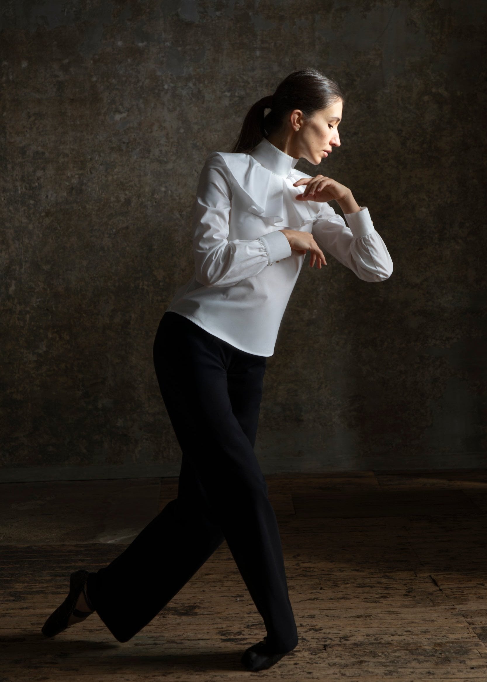 Une femme portant le chemisier Giselle blanc avec un col plissé de Sidonie Paris en pantalon noir prend une pose de danse contemporaine sur un fond texturé. Ses cheveux sont attachés en arrière tandis qu'elle étend gracieusement un bras vers l'avant, capturant la fluidité et l'élégance qui rappellent la poésie dans l'art.