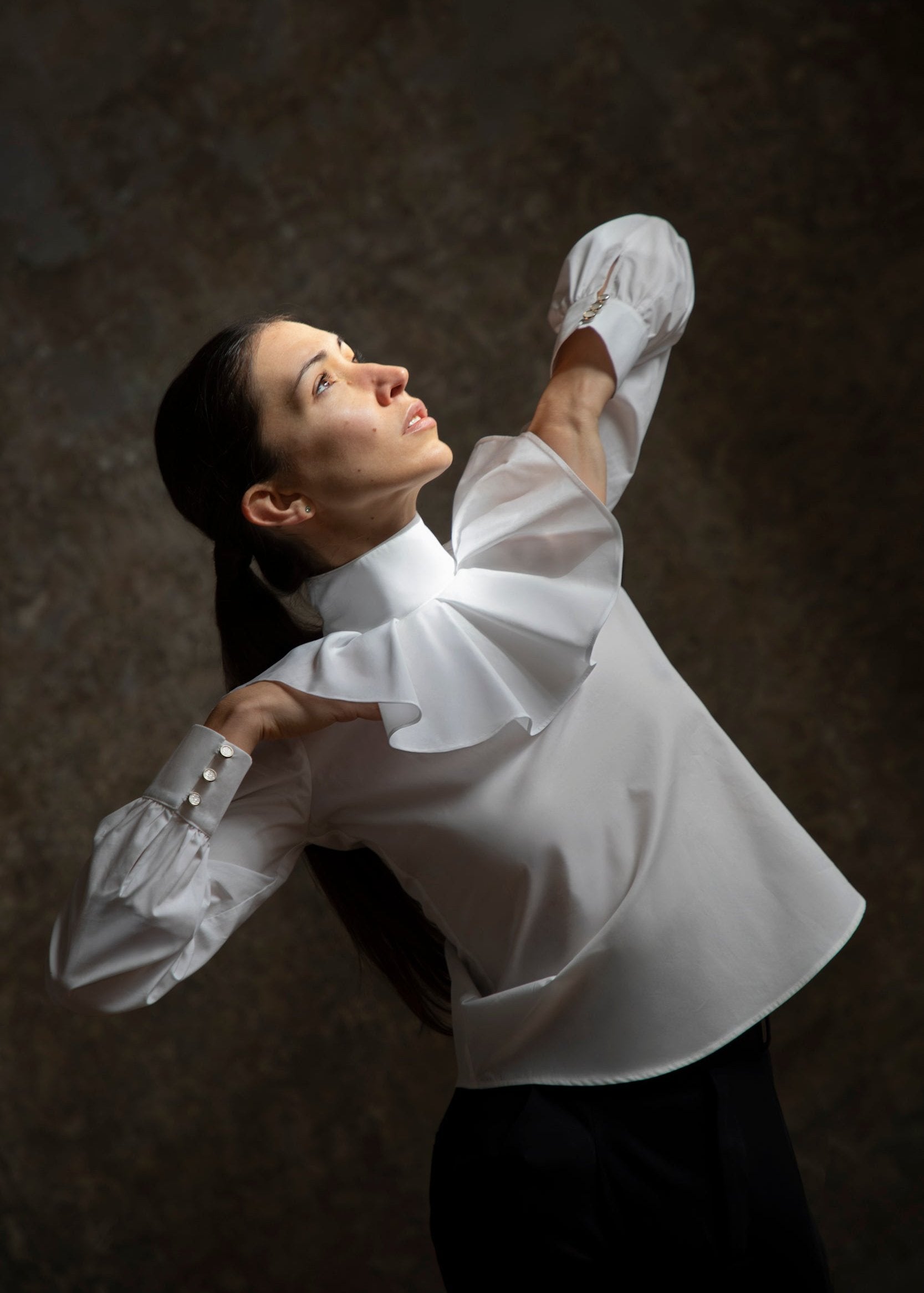 Une personne adopte une pose artistique, les bras levés, portant l'élégant Chemisier Giselle : une blouse blanche ornée d’un grand col plissé signée Sidonie Paris. En arrière-plan, une surface sombre et texturée met en valeur la lumière qui accentue son visage et sa tenue, capturant ainsi l’essence d’une véritable œuvre d’art.