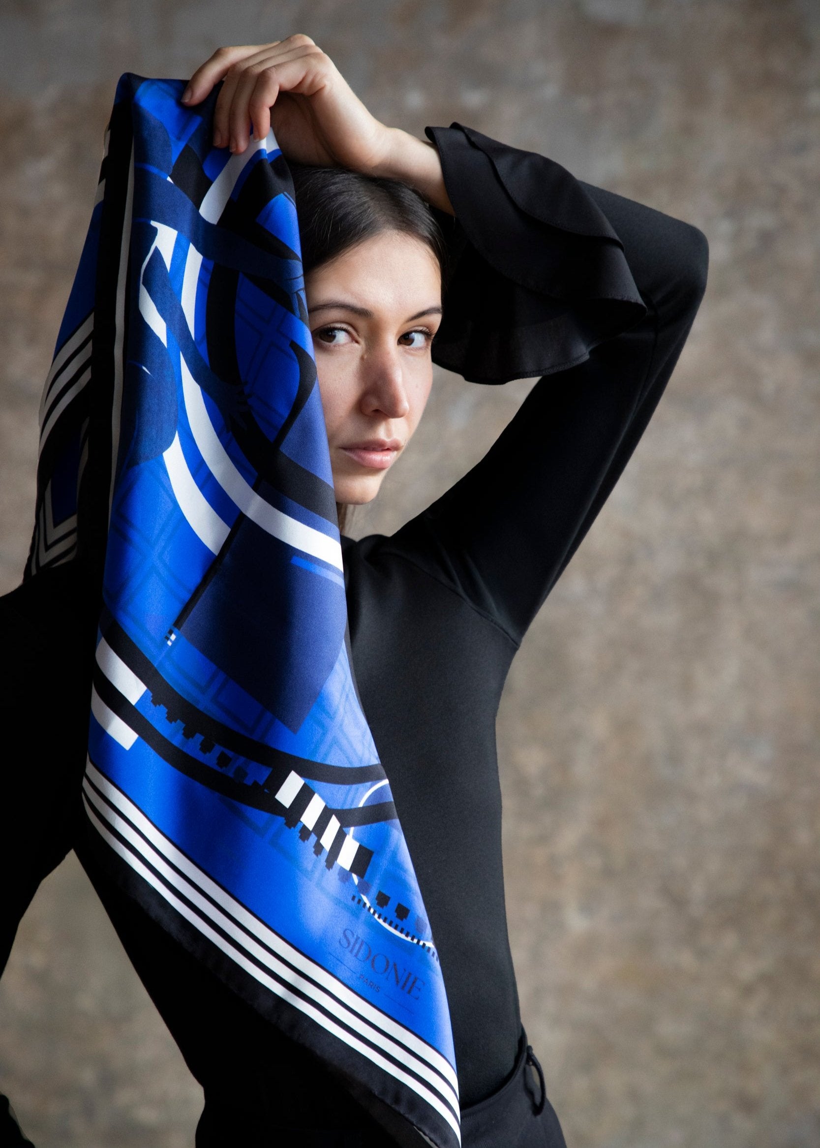 Une femme tient un foulard en soie bleu avec des motifs graphiques, en le drapant élégamment sur son épaule. Elle porte un haut noir à manches évasées et regarde intensément l'objectif. L'arrière-plan est flou, créant une atmosphère artistique et sophistiquée, mettant en valeur le foulard et le style de la modèle.