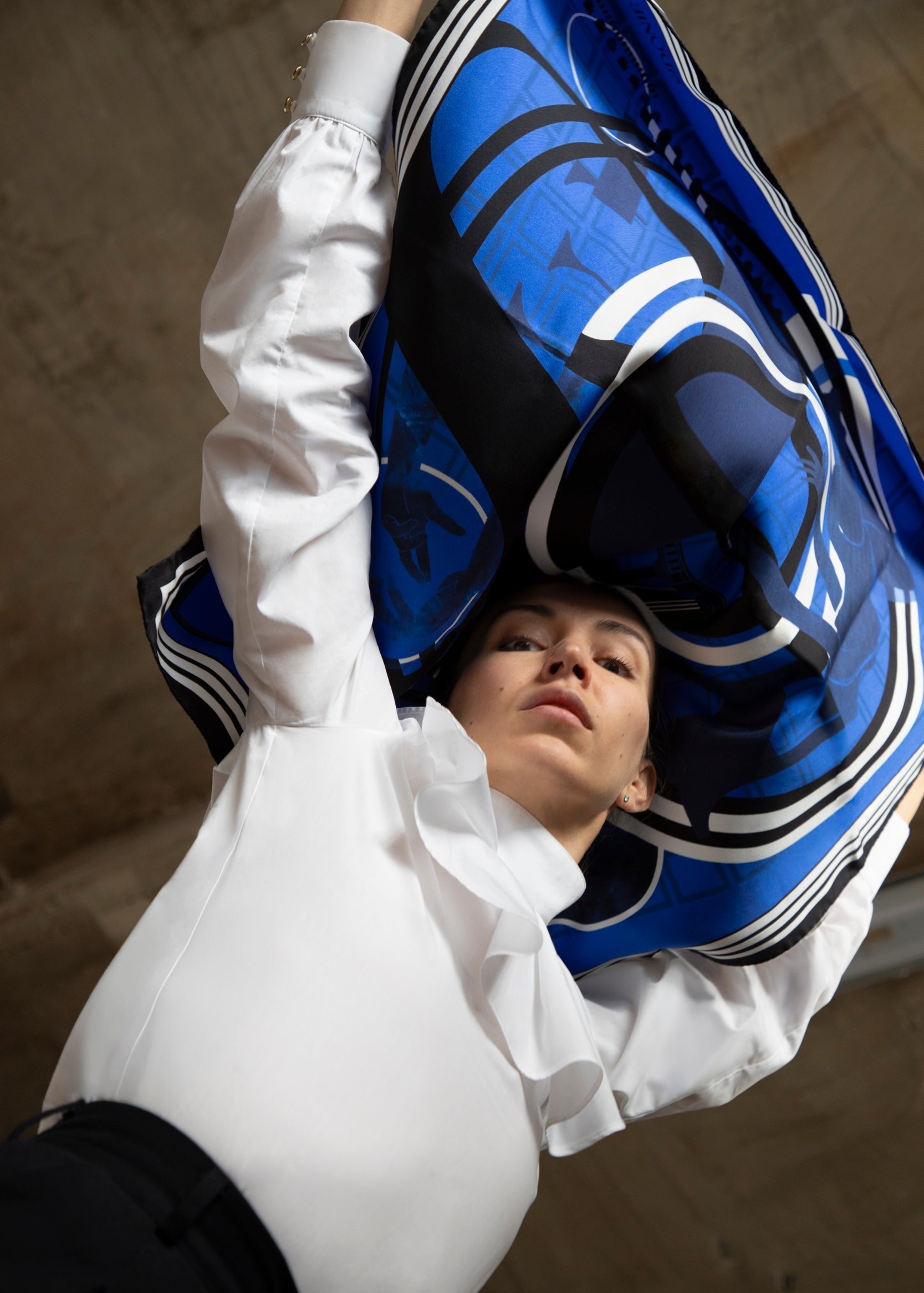 Un modèle portant un foulard en soie bleu orné de motifs graphiques, levé au-dessus de sa tête, avec une chemise blanche à col froncé et un pantalon noir. L'image est prise en perspective, mettant en valeur le foulard qui flotte et la concentration du modèle, le fond étant flou pour accentuer le sujet principal.