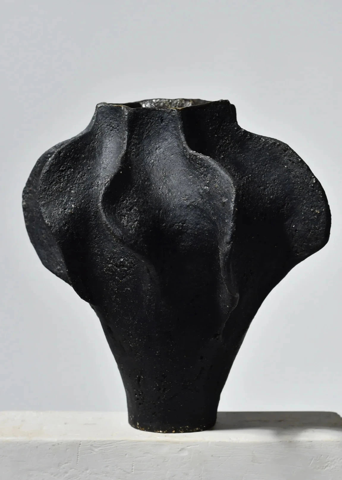 Vase en grès noir, présentant une forme unique avec des contours ondulés et texturés, idéal comme objet d'art décoratif. Le contraste entre la surface mate et la lumière crée une esthétique moderne et élégante, parfaite pour embellir tout intérieur.